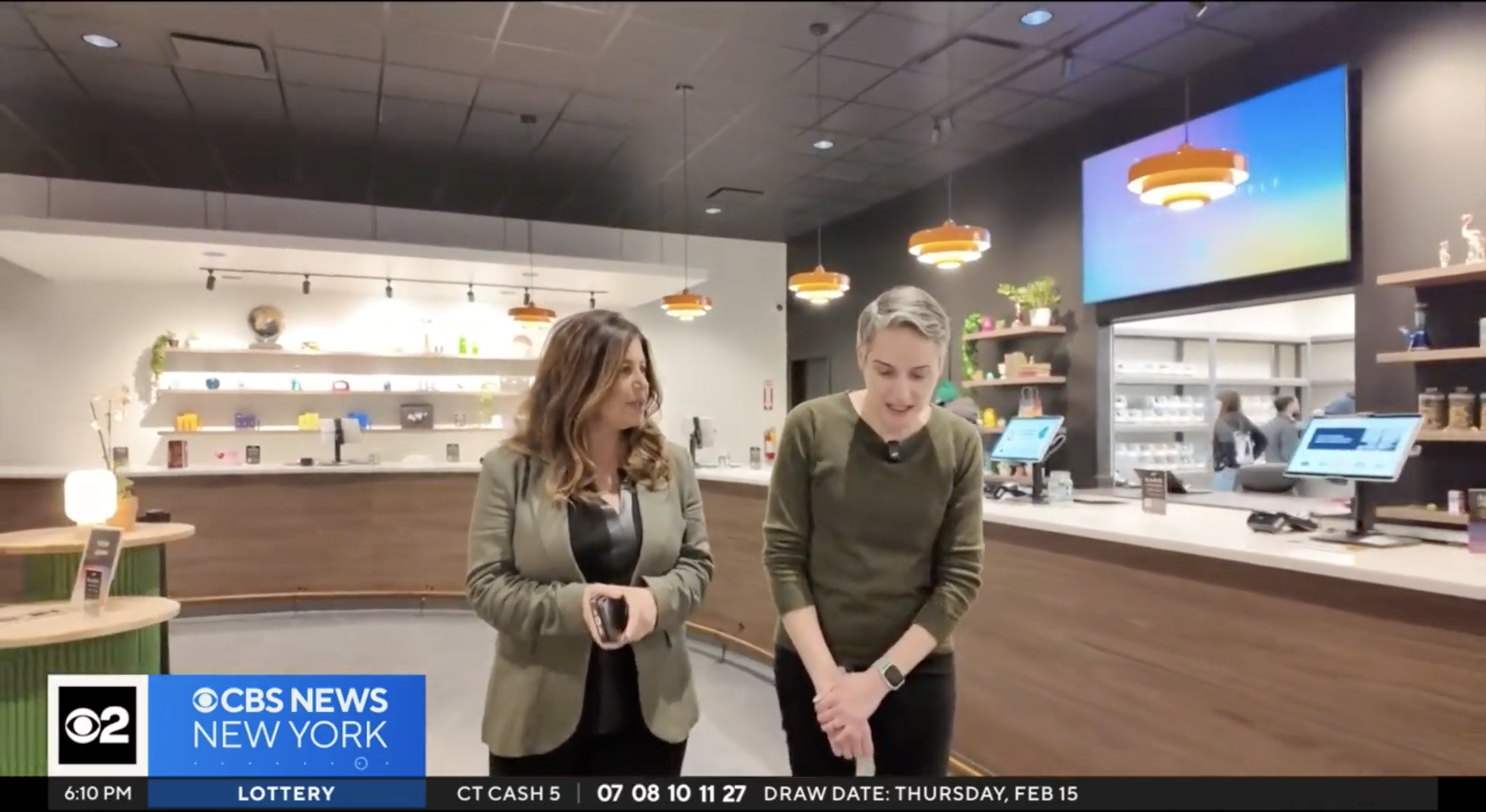 Two women are standing and talking inside a modern retail space with wooden counters and shelves displaying various items. The woman on the left has long wavy hair and is wearing a green blazer over a black top, while the woman on the right has short gray hair and is wearing a green sweater and a smartwatch. In the background, there are hanging orange pendant lights, a large screen, and a few people working behind a counter with computer monitors. The CBS News New York logo and a lottery draw date are visible in a lower-third graphic at the bottom of the image.